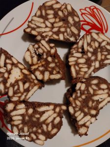 nycsiklandó, házi készítésű kekszszalámi egy fa vágódeszkán tálalva. A sötétbarna, kakaós masszába ágyazott világos háztartási kekszdarabok márványos mintát adnak a szeleteknek. A rudak külseje hófehér kókuszreszelékkel van gazdagon megszórva, a háttérben pedig néhány egész keksz és egy csésze gőzölgő kávé látható.