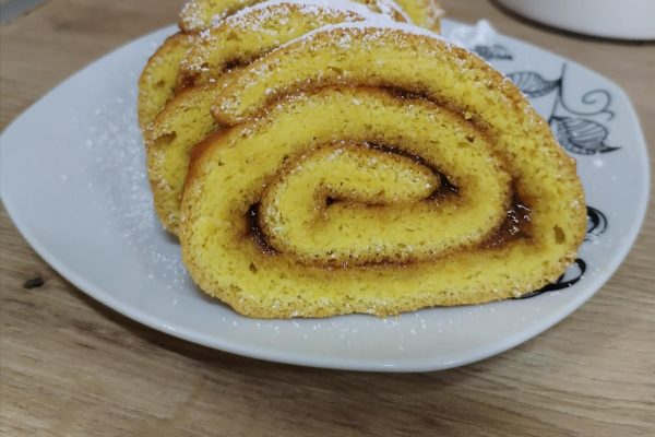 Egy fehér kistányéron, porcukorral meghintve, öt szelet, baracklekvárral töltött piskótatekercs látható, fa felületen elhelyezve, a háttérben egy konyhai kendővel.