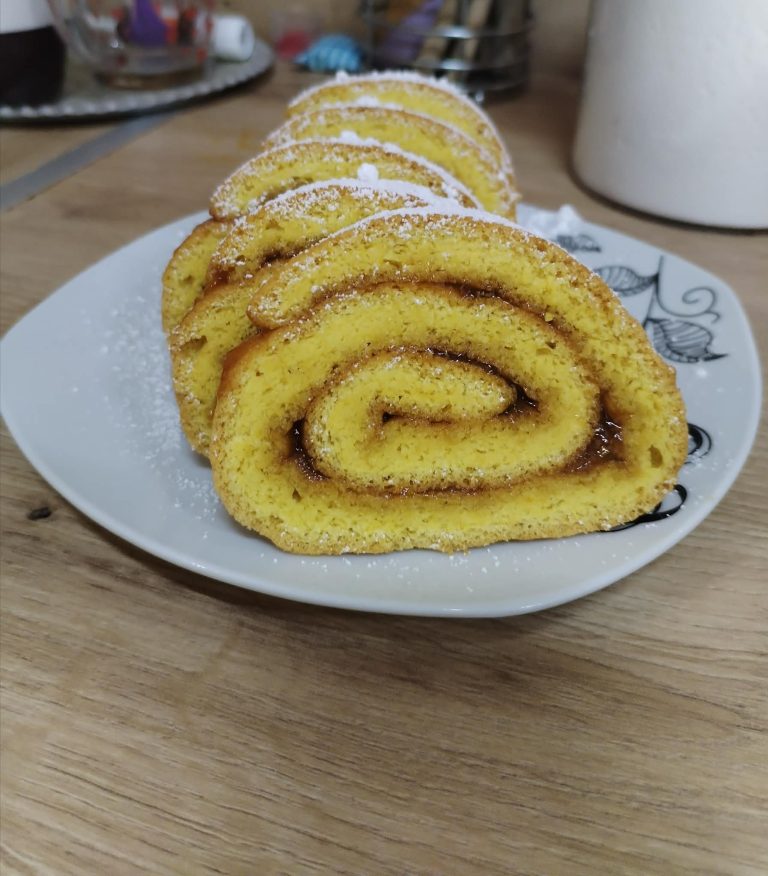 Egy fehér kistányéron, porcukorral meghintve, öt szelet, baracklekvárral töltött piskótatekercs látható, fa felületen elhelyezve, a háttérben egy konyhai kendővel.