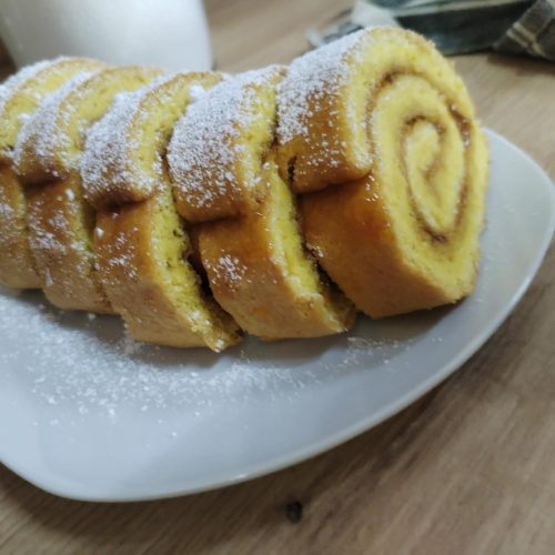 Egy fehér kistányéron, porcukorral meghintve, öt szelet, baracklekvárral töltött piskótatekercs látható, fa felületen elhelyezve, a háttérben egy konyhai kendővel.